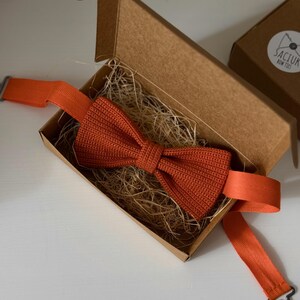 May include: An orange bow tie with a textured knit design, presented in an open cardboard box filled with straw. The bow tie has an adjustable strap and metal clasp. A second box with a logo is visible in the background.