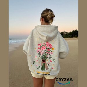 May include: A light gray hoodie with a floral bouquet design on the back. The bouquet features pink and yellow flowers with green leaves. The text "GIFT GIVING" is printed below the design. The hoodie is worn on a beach.