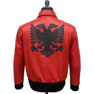 Op de afbeelding: Rode leren jas met een zwart dubbelkoppig adelaarsymbool op de rug. De jas heeft een zwarte geribbelde kraag, manchetten en zoom. De jas wordt op een mannequin getoond.