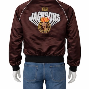 Könnte beinhalten: Braune Satin-Bomberjacke mit dem weißen Schriftzug "JACKSONS" und einem Logo mit einer Fackel und den Worten "WORLD TOUR 84-85". Die Jacke hat schwarze Bündchen, Kragen und Saum sowie weiße Paspeln.