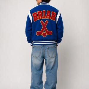May include: A blue varsity jacket with white striped ribbed cuffs and collar, featuring the text BRIAR in red and yellow block letters above a graphic of two crossed hockey sticks in red and yellow on the back, paired with light blue denim jeans.