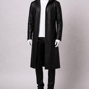 Puede incluir: Un abrigo largo negro de cuero con capucha, usado sobre una camiseta blanca y pantalones negros. El maniqu&iacute; lleva zapatillas grises. El abrigo tiene un dise&ntilde;o elegante y minimalista.