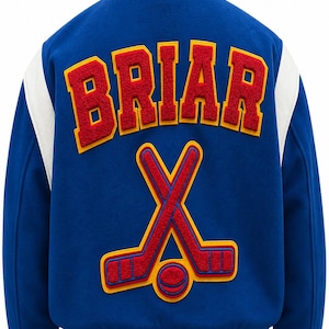 Könnte beinhalten: Blaue Collegejacke mit weißen Ärmeln und Besatz. Das Wort "BRIAR" ist rot mit gelbem Rand auf dem Rücken gestickt. Darunter gekreuzte Hockeyschläger und ein Puck.