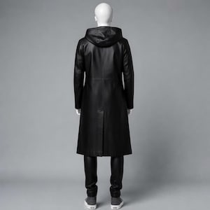 Puede incluir: Un abrigo largo de cuero negro con capucha, exhibido en un maniqu&iacute;. El abrigo llega hasta los tobillos y tiene un corte entallado. El maniqu&iacute; tambi&eacute;n lleva pantalones negros y zapatillas grises.