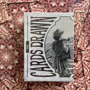 Op de afbeelding: Een kaartspel met een decoratief ontwerp. De kaartenbox heeft de tekst "THE CARDS DRAWN" in een verticale lay-out, met een zwart-wit illustratie van een persoon die naar een landschap kijkt. De kaarten zijn gerangschikt op een patroon oppervlak.