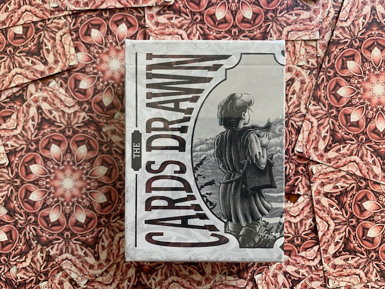 Op de afbeelding: Een kaartspel met een decoratief ontwerp. De kaartenbox heeft de tekst "THE CARDS DRAWN" in een verticale lay-out, met een zwart-wit illustratie van een persoon die naar een landschap kijkt. De kaarten zijn gerangschikt op een patroon oppervlak.