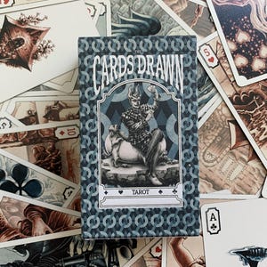 以下が含まれることがあります： 青と白の模様の箱に入ったタロットカードのデッキ。「The Cards Drawn Tarot」と書かれた箱には、馬に乗った人物のイラストが描かれています。さまざまなイラストやカードのマークが、箱の周りに散らばっています。