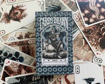 Op de afbeelding: Een tarotkaartspel met een blauw en wit patroon doos. De doos leest "The Cards Drawn Tarot" en heeft een illustratie van een figuur op een paard. De kaarten zijn verspreid rond de doos, met verschillende illustraties en kaartspellen.
