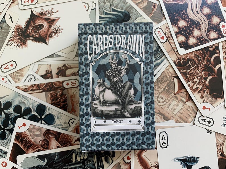 以下が含まれることがあります： 青と白の模様の箱に入ったタロットカードのデッキ。「The Cards Drawn Tarot」と書かれた箱には、馬に乗った人物のイラストが描かれています。さまざまなイラストやカードのマークが、箱の周りに散らばっています。