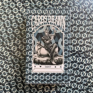 以下が含まれることがあります： 青と白の幾何学模様のタロットカードのデッキ。カードボックスには「THE CARDS DRAWN」の文字と、動物の上に座っている人物のイラストが描かれています。「TAROT」の文字がボックスに印刷されています。