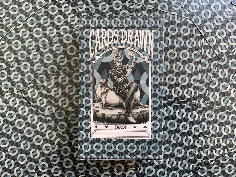 以下が含まれることがあります： 青と白の幾何学模様のタロットカードのデッキ。カードボックスには「THE CARDS DRAWN」の文字と、動物の上に座っている人物のイラストが描かれています。「TAROT」の文字がボックスに印刷されています。