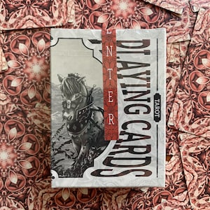 Op de afbeelding: Een kaartspel "Playing Cards Tarot" met een zwart-wit illustratie van een paard op de voorkant. Het spel is verpakt met een rode strip met de tekst "Enter". De achtergrond toont verspreide kaarten met een rood-wit bloemenpatroon.