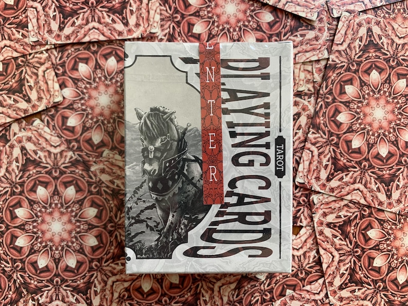 Op de afbeelding: Een kaartspel "Playing Cards Tarot" met een zwart-wit illustratie van een paard op de voorkant. Het spel is verpakt met een rode strip met de tekst "Enter". De achtergrond toont verspreide kaarten met een rood-wit bloemenpatroon.