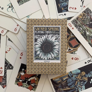 以下が含まれることがあります： 青と金の星の模様が繰り返されるベージュの箱に入ったSacred Springトランプのデッキ。中央にはヒマワリのイラストと「Sacred Spring Playing Cards by The Cards Drawn MMXXVI」という文字が描かれており、散らばったトランプの上に置かれています。