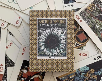 Op de afbeelding: Een kaartspel Sacred Spring in een beige doos met een herhalend blauw en goud sterrenpatroon, met een centrale illustratie van een zonnebloem en de tekst Sacred Spring Playing Cards by The Cards Drawn MMXXVI, bovenop verspreide speelkaarten.