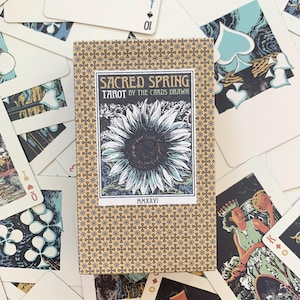 以下が含まれることがあります： Sacred Spring Tarot by The Cards Drawnの長方形のタロットカードデッキ。青と茶色の星模様が繰り返されるタンカラーの箱に入っています。中央のラベルにはヒマワリのイラストと「Sacred Spring Tarot MMXXVI」という文字が描かれています。