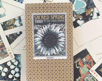 Op de afbeelding: Een rechthoekig kaartspel Sacred Spring Tarot by The Cards Drawn, in een beige doos met een herhalend blauw en bruin sterpatroon. Het centrale label toont een illustratie van een zonnebloem met de tekst Sacred Spring Tarot MMXXVI.