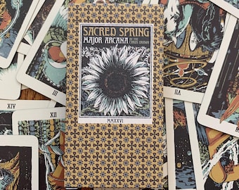 Op de afbeelding: Een rechthoekig kaartendoosje voor tarotkaarten met een beige achtergrond en een herhalend donkerblauw sterrenpatroon. Het centrale label toont een zwart-witte zonnebloemillustratie met de tekst Sacred Spring Major Arcana en MMXXVI.