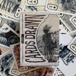 Op de afbeelding: Een kaartspel met de titel "The Cards Drawn" met een grijswaardenillustratie van een persoon. De kaarten zijn verspreid rond de doos en tonen verschillende kleuren en getallen. De algehele esthetiek is vintage en artistiek.