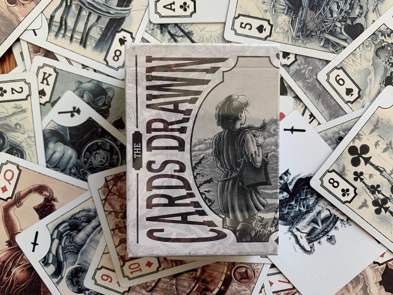 Op de afbeelding: Een kaartspel met de titel "The Cards Drawn" met een grijswaardenillustratie van een persoon. De kaarten zijn verspreid rond de doos en tonen verschillende kleuren en getallen. De algehele esthetiek is vintage en artistiek.