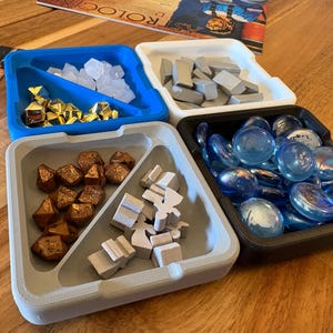 May include: Four divided trays filled with game pieces. The blue tray holds clear and gold pieces, the white tray has grey pieces, the grey tray contains brown and white pieces, and the black tray is filled with blue glass stones. A game board is in the background.