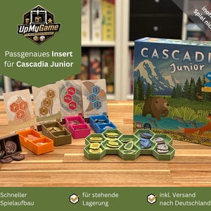 Könnte beinhalten: Ein Spielkarton mit der Aufschrift "Cascadia Junior" steht neben verschiedenen Spielkomponenten, darunter farbige Kunststoffschalen und sechseckige Spielsteine. Die Box zeigt Illustrationen eines Bären und eines Fuchses in einer natürlichen Umgebung. Der Text zeigt 6+.
