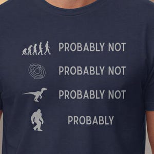 May include: Navy blue t-shirt with a humorous graphic. The design features a progression of figures, a maze, a dinosaur, and a Bigfoot, each paired with "PROBABLY NOT," except for Bigfoot, which says "PROBABLY."