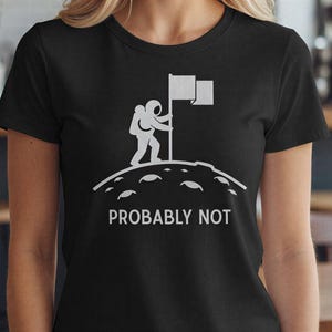 May include: Black t-shirt featuring a white graphic of an astronaut planting a flag on a planet's surface. The text "PROBABLY NOT" is printed below the graphic. The shirt is made of a soft material.