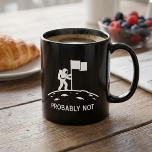 May include: Black coffee mug with a white graphic of an astronaut planting a flag on a planet, with the words "PROBABLY NOT" below. The mug is on a wooden table with a croissant and berries.