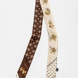 May include: A long, narrow silk scarf featuring a dual-sided design. One side is dark brown with a repeating white geometric pattern and a floral motif with the text "Soft Bloom Floral Whisper". The other side is cream with floral illustrations, paisley, and a black polka dot section.