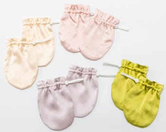 May include: Four pairs of baby mittens in satin fabric, each featuring a gathered elastic wrist with a white drawstring. The colors include cream, light pink, lavender, and lime green, arranged in a cluster on a plain white background.