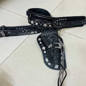 Pode incluir: Um conjunto de cinto e coldre de couro preto. O cinto apresenta uma fivela prateada e tachas decorativas prateadas. O coldre é gravado com desenhos florais e tem uma fivela prateada. O conjunto é projetado para um revólver de ação simples.
