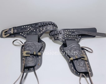 May include: Black leather gun belt and holster set. The set features embossed floral designs, silver-colored studs, and brass-colored buckles. The holsters have a strap with a buckle to secure the gun.