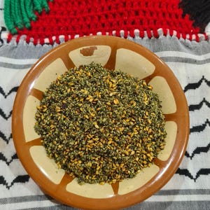 Peut inclure: Un bol rond en céramique marron rempli d'un mélange d'herbes vertes et de graines de sésame dorées. Le bol est posé sur un tissu à motifs noir et blanc, avec un fond crocheté rouge, blanc, vert et noir.