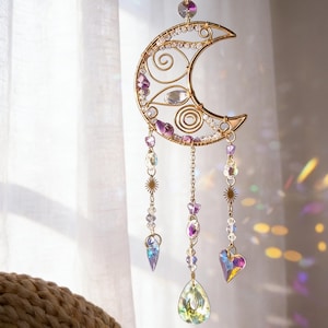 Op de afbeelding: Een goudkleurige maansvanger met heldere en paarse kristallen. De maan heeft een draadframe met decoratieve krullen en hangende kettingen met kristallen, sterren en een hartvormige bedel. Het hangt voor een wit gordijn.