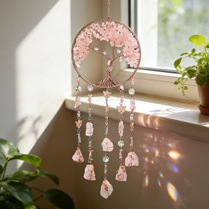 May include: A rose gold metal tree of life dream catcher with pink crystal accents. The circular design features a detailed tree with pink crystal leaves and hanging strands of crystals, butterflies, and raw rose quartz stones.