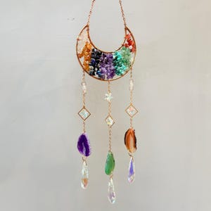 Pu&ograve; includere: Un acchiappasole a forma di mezzaluna con una cornice color rame, riempito di pietre preziose colorate. Presenta catene pendenti con cristalli, ciondoli a forma di stella e fette di agata viola, verde e marrone. Un elemento decorativo.