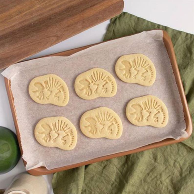 May include: Six light-colored, dinosaur-shaped cookies on parchment paper inside a brown baking pan. The cookies have a detailed imprint of a dinosaur. A wooden cutting board and green fabric are in the background.