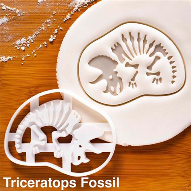 May include: A white plastic Triceratops fossil cookie cutter next to a white dough imprint of the same design. The cutter is on a wooden surface with flour sprinkled around the dough. The text "Triceratops Fossil" is at the bottom.