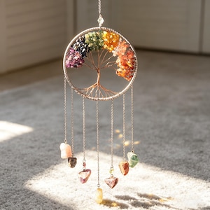 May include: A handcrafted tree of life wall hanging. The design features a circular silver-colored frame with a wire tree and colorful gemstones representing the chakras. Dangling chains hold various gemstone pendants. The piece is designed to be hung.