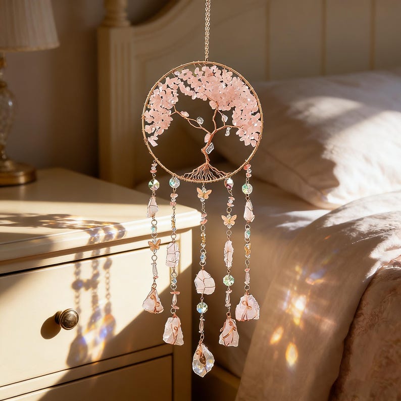 Puede incluir: Un atrapasue&ntilde;os decorativo con un marco de metal dorado rosa con un dise&ntilde;o de &aacute;rbol. El &aacute;rbol est&aacute; adornado con cristales rosas, y los hilos colgantes sostienen cristales, cuentas y dijes de mariposa. La paleta de colores es rosa suave y dorado.