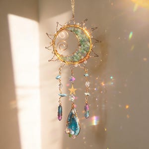 Pode incluir: Um apanhador de sonhos com tema celestial com uma estrutura de arame dourado em um design de lua crescente e explosão solar. A lua é preenchida com pedras verdes, e penduradas nela estão contas, cristais e um pingente em forma de gota. Uma estrela dourada também está incluída.