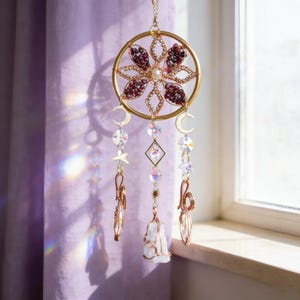May include: A gold-colored dream catcher with a floral design, adorned with red gemstones and a central pearl. It features hanging crystals, moons, stars, and a white crystal pendant, creating a celestial theme. The dream catcher hangs in front of a purple curtain.