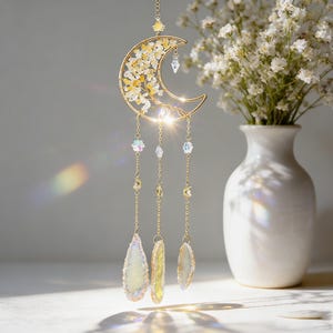 Puede incluir: Un atrapasoles dorado en forma de media luna con detalles de cristal amarillo y transparente. El atrapasoles tiene colgantes de cristal y piedra. Un jarrón blanco con flores blancas está en el fondo.