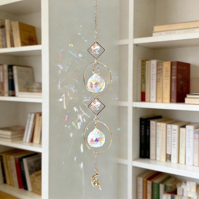 May include: A hanging suncatcher with a gold chain, geometric shapes, and crystal spheres. The design includes a diamond, a circle, and a crescent moon charm. The crystals refract light, creating a rainbow effect.