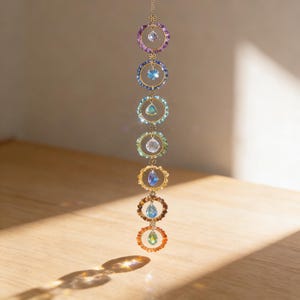 May include: A decorative hanging ornament with seven crystal pendants in a range of colours. Each crystal is set within a gold-coloured ring. The colours include purple, blue, green, and orange. The ornament is designed to reflect light.