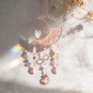 May include: A gold-coloured metal crescent moon-shaped suncatcher. The moon is filled with pink crystals and adorned with hanging crystals and gold-coloured accents. A clear crystal is attached to the top of the moon. The suncatcher is hanging from a chain.