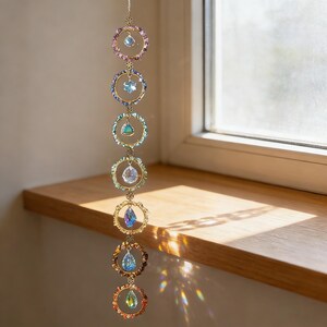 May include: A decorative suncatcher featuring seven circular elements in a range of colours, each with a crystal pendant. The rings are linked by a fine gold chain, creating a beautiful display of light and colour. The suncatcher is designed to catch and reflect sunlight.