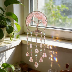 Puede incluir: Un atrapasue&ntilde;os de color oro rosa con un dise&ntilde;o de &aacute;rbol, lleno de cristales rosas. El atrapasue&ntilde;os tiene hilos colgantes de cristales, cuentas y dijes de mariposas. El dise&ntilde;o est&aacute; colgado frente a una ventana.