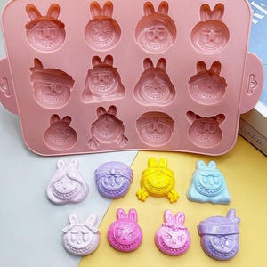 May include: A pink silicone mold with 12 bunny-faced cavities. Several colorful bunny-shaped candies are displayed below the mold. The candies are various colors, including pink, purple, yellow, and blue. The mold has handles on each side.